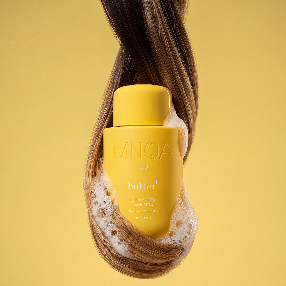 A yellow bottle of MNQA by 9 butter+ shampoo is wrapped in flowing brown hair and white foam against a solid yellow background.