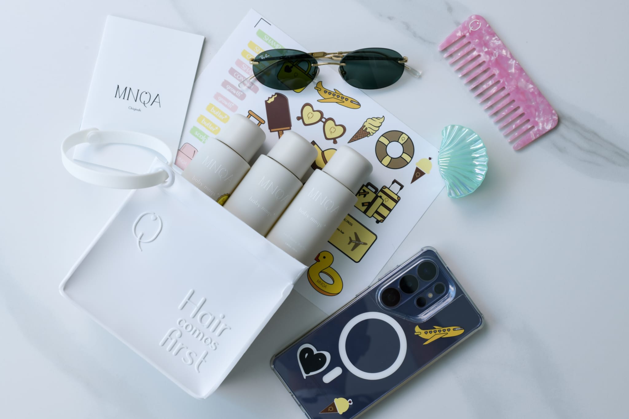 A styled flat-lay of hair care and beauty accessories on a light marble surface, including a white box with embossed lettering, three white cylindrical bottles, a pink comb, green sunglasses, a seashell-shaped hair tie, a printed sticker sheet, and a dark phone case.